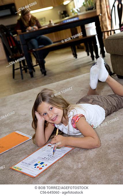 smiling young girl does her homework on floor, mother in background