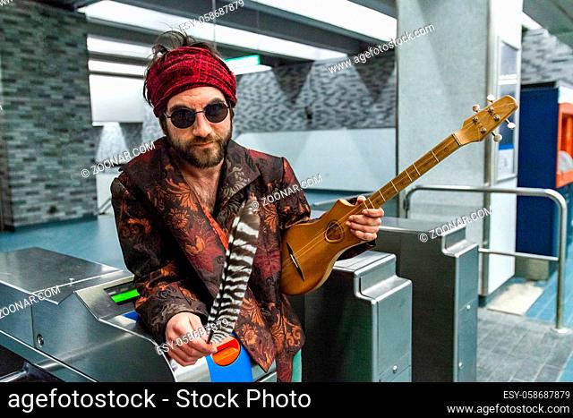 A portrait of a medicine man holding sacred eagle feather and wooden guitar, as he leans against the ticket entry gates of a subway station, wearing bandanna