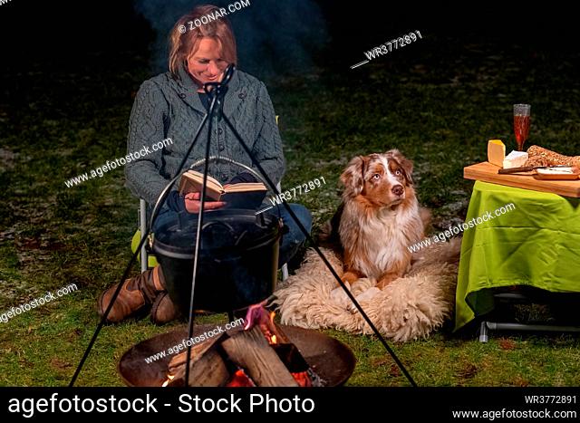 Australian Shepherd dog is lying by the campfire, the kettle with food is cooking and steaming, The woman is reading a book