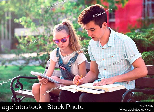 Students making the notes learning from books sitting on a bench in a park. Young boy wearing a blue shirt and dark jeans