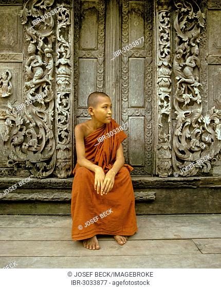 Monk sitting in front of a wooden door with teak carvings, Shwe In Bin Kyaung Monastery