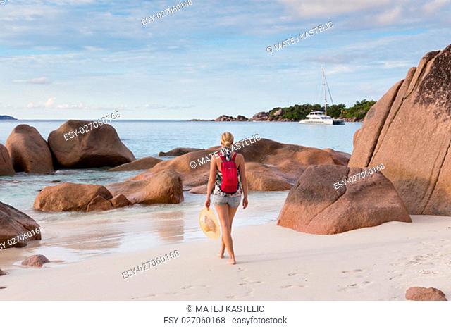 Woman wearing smal backpack, scarf, jeans shorts and beach hat, enjoying view of Anse Lazio beach on Praslin Island, Seychelles