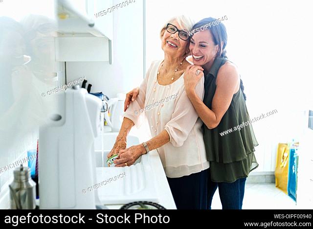 Cheerful granddaughter hugging grandmother washing dishes in kitchen at home