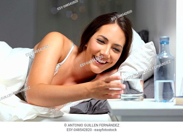 Happy woman reaching a glass of water lying on the bed in the night at home