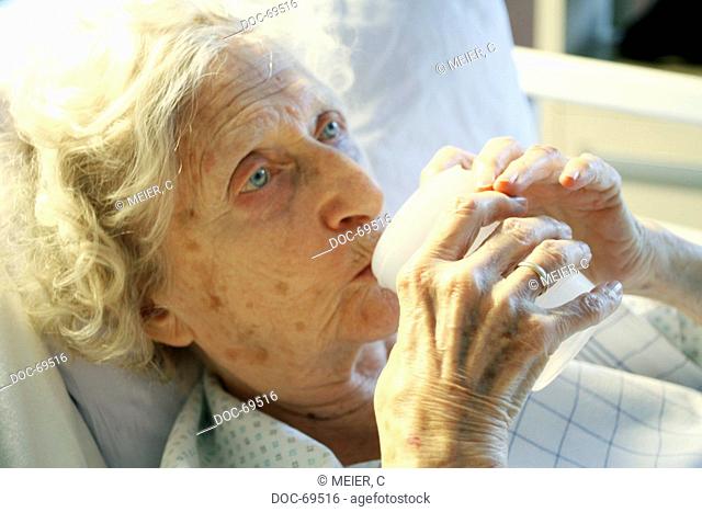 Elderly woman is lying in a bed and drinks from a mug