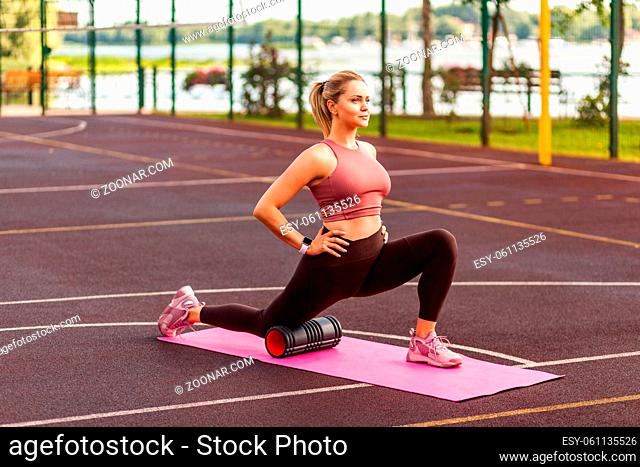 Sporty fit girl training on mat outdoor summer day, using foam roller massager to support knee while stretching legs muscles, doing fascia exercise