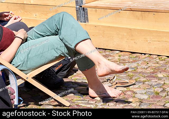 11 August 2023, Lower Saxony, Lüneburg: A woman with rolled-up pants and bare feet enjoys the sun in a deck chair on the market square on this warm summer day