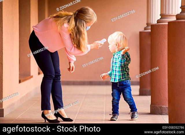 Young preschool teacher in medical mask checking kid's temperature with temporal thermometer. Every kid needs to be checked before entering kindergarten or...