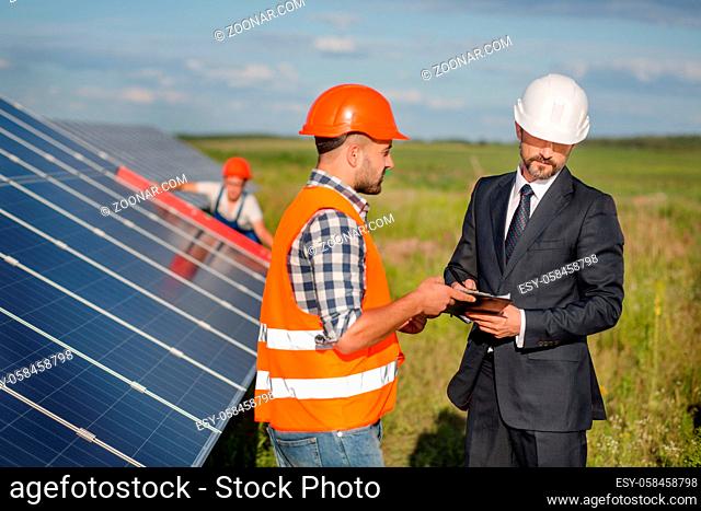 Businessman signing contract with foreman, technician examining solar panels at backdrop. Signing of agreement at solar energy station in the field