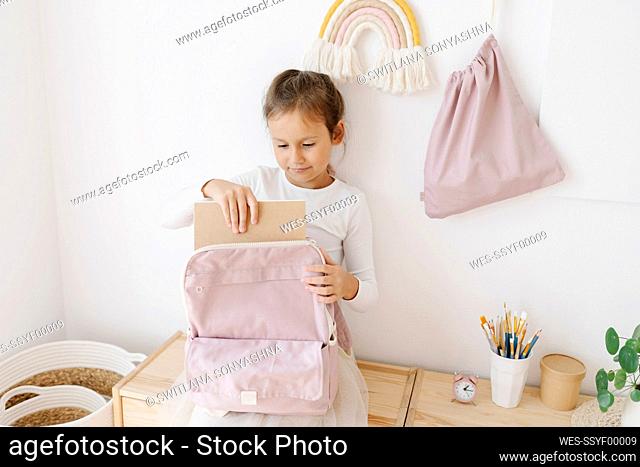 Cute girl removing book from backpack sitting on table