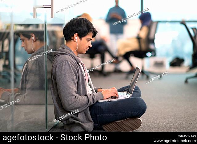 young indian software developer man using laptop computer writing programming code while sitting on the floor at modern creative startup office