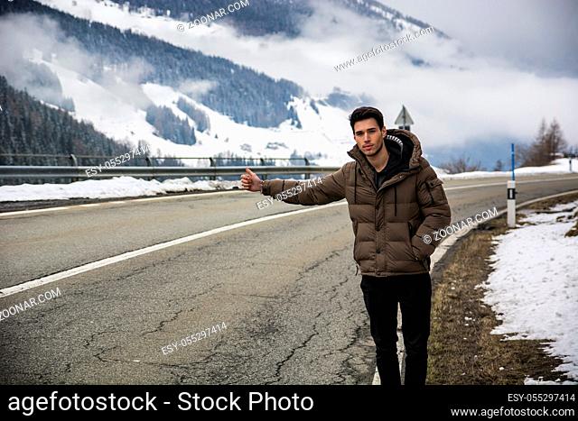 Young man thumbing a ride on roadside. Snowy mountain on background