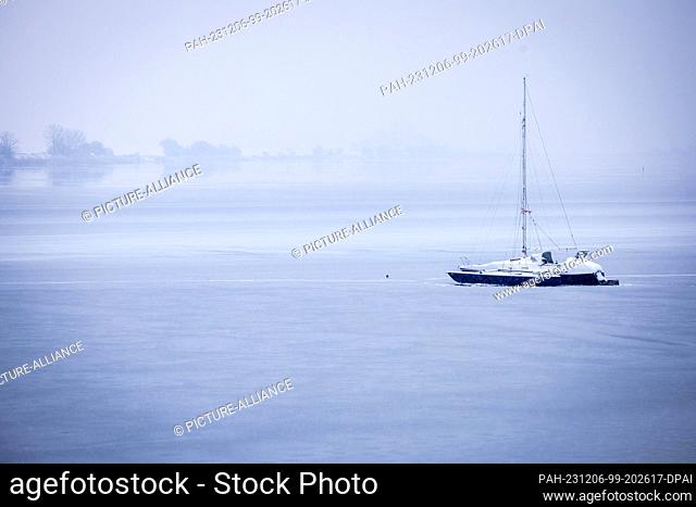 06 December 2023, Mecklenburg-Western Pomerania, Kirchdorf (poel): A small sailing boat covered in snow is frozen in thin ice in front of the harbor on the...