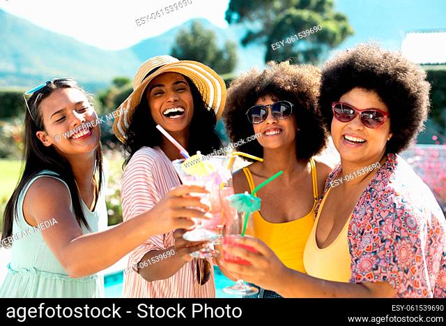 Cheerful biracial female friends toasting cocktails and posing while enjoying pool party in summer