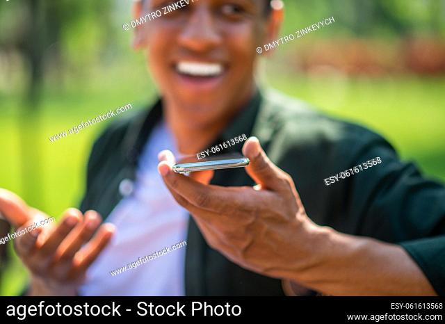 Audio message. Smartphone in hand of dark-skinned man gesturing happily talking in park on warm day