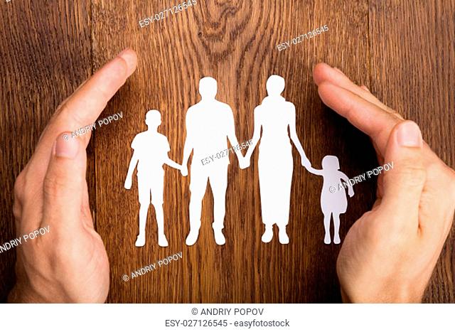 Close-up Of Person Hand Protecting Family Papercut At Wooden Desk