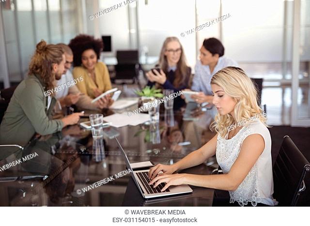 Young businesswoman using laptop on conference table with team in background