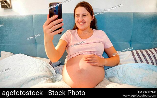 Happy smiling pregnant woman making selfie on smartphone before going to bed at night