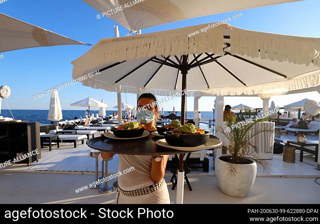23 June 2020, Spain, Palma: A waitress with a face mask carries plates with food on a tray in the bar Purobeach in Cala Estancia on the beach Playa de Palma