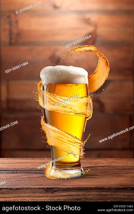 Creative flying spiral yellow splash aroung full glass of fresh cold light beer on a wooden table against wall from wood, copy space