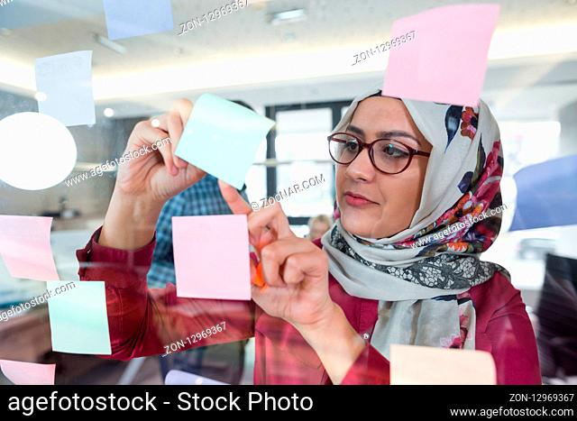 Beautiful Arab woman wearing hijab putting sticky notes on glass in modern office