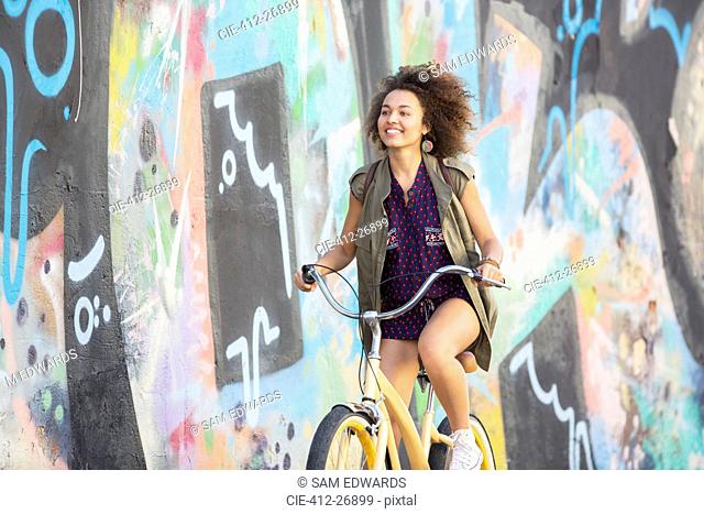 Smiling brunette woman riding bicycle along urban multicolor graffiti wall