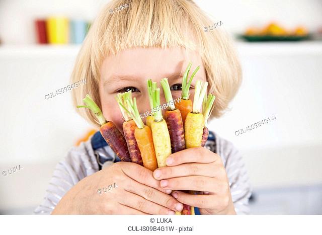 Portrait of cute girl in kitchen holding bunch of colourful carrots to her face