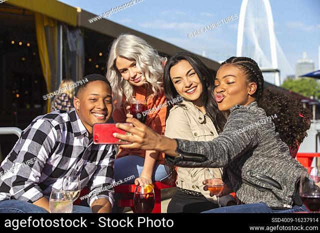 Group of young co-workers hanging out on outdoor patio having drinks for happy hour, woman taking group photo with mobile phone