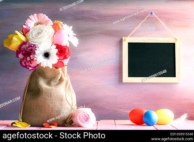 Easter banner with a bouquet of flowers in a jute sack, painted eggs and a blank blackboard hanging on a purple wooden background, on a sunny day
