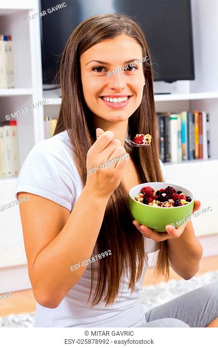 Happy young woman eating cereal muesli with fruits in bowl at home. Healthy Food and Dieting concept