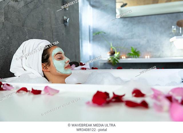 Side view of a young Caucasian brunette woman lying in a bath with lit candles and rose petals on the side, leaning back with her hair wrapped in a towel and...