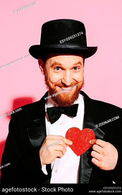 Smiling man in a tuxedo and top hat holding a red heart
