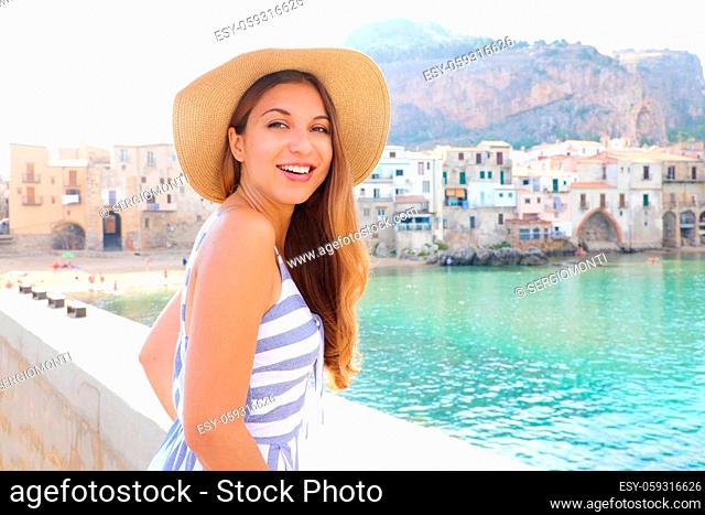 Portrait of attractive smiling latin woman with hat and summer dress looking at camera with Cefalu village on the background, Sicily Island, italy