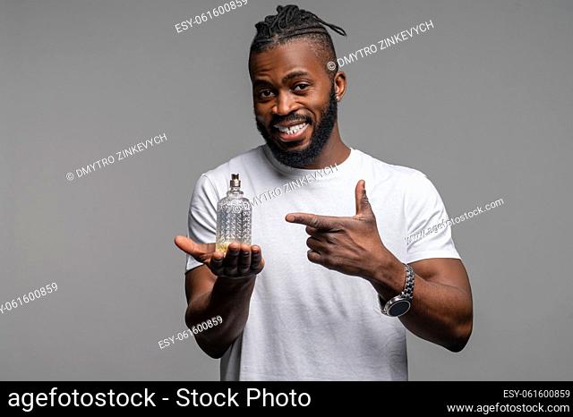 Waist-up portrait of a cheerful guy pointing at a small glass perfume bottle in his hand