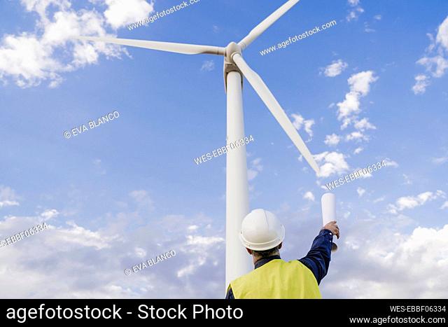 Engineer with blueprint looking at wind turbine by sky