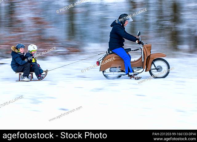 03 December 2023, Mecklenburg-Western Pomerania, Pokrent: A family is out and about in the snow on a moped with a sledge attached (shot with longer exposure...