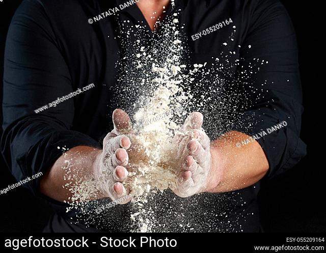 chef in black uniform sprinkles white wheat flour in different directions, product scatters dust, black background, close up
