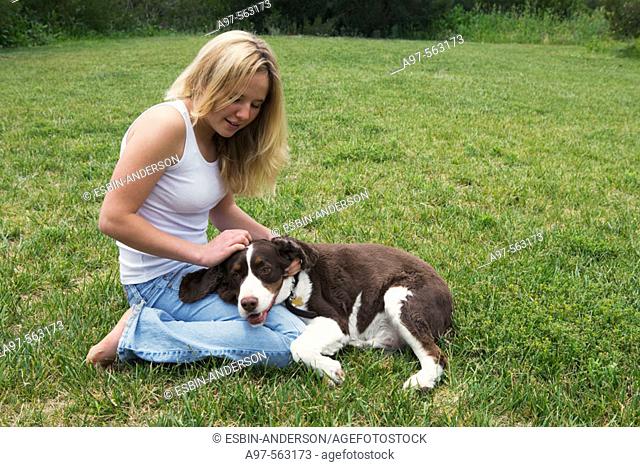 Smiling blonde teen girl in blue jeans and white tank top kneeling on grass lawn, pets her dog's head resting on her leg