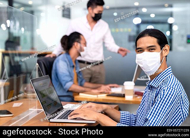 Asian Office employee with protective face mask working in new normal office with social distance practice to his colleague in background prevent coronavirus...