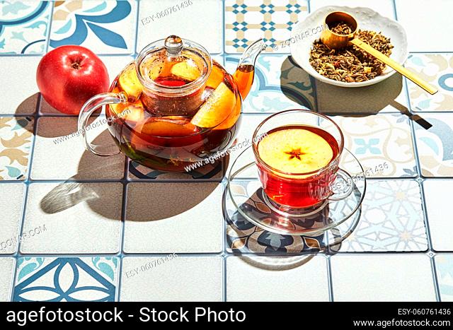 Fruit tea with apples and thyme in glass teapot and cup on table made of colored tiles with hard shadows