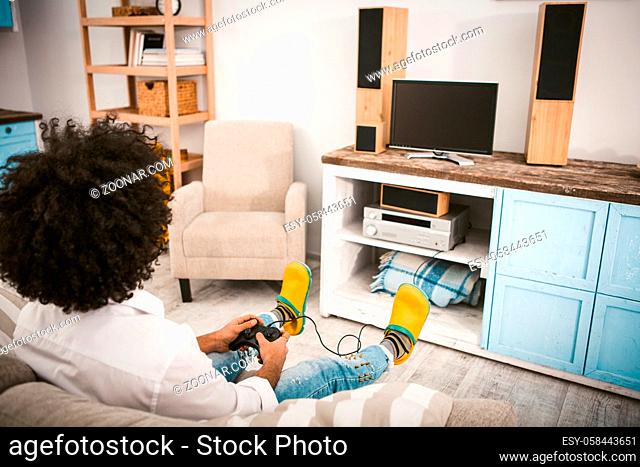 Stylish guy plays computer games sitting on couch. Young unrecognizable man with afro hairstyle in yellow slippers relaxing in home interior