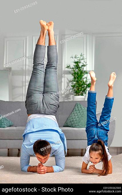 father copy imitate active child girl doing gymnastic handstand exercise at home, dad and daughter stand on hands, sporty family