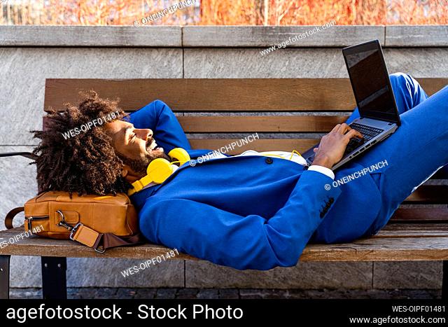 Happy businessman with headphones using laptop by lying down on bench