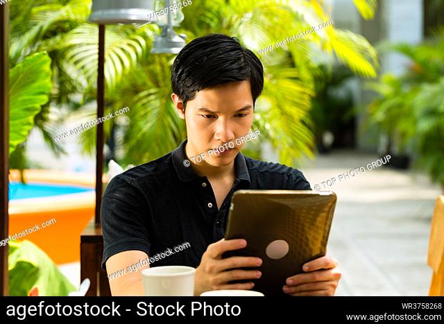 Asian man is sitting in a bar or cafe outdoor and is surfing the internet with a tablet computer