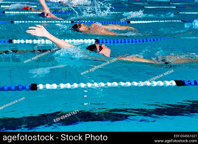 Side view of multi-ethnic group of male swimmers at swimming pool, racing each other in lanes, swimming backstroke. Swimmers training hard for competition