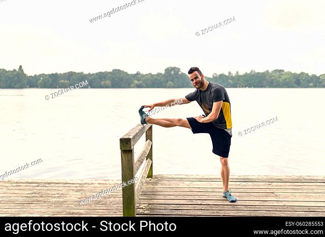 Young man doing stretching exercises on a wooden deck overlooking a lake raising his leg on a fence rail as he smiles at the camera in a healthy lifestyle...