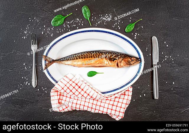 Whole smoked mackerel fish on white oval plate, gray black stone like table under, rock salt and green leaves salad near. View from above