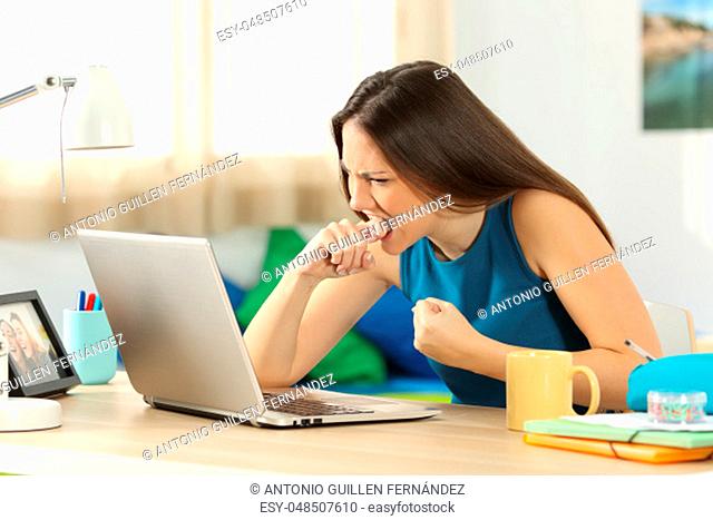 Angry student with a laptop failure sitting in a desk in her room in a house interior with a window in the background