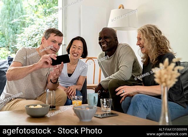 Senior man sharing pictures on smart phone with friends while sitting on sofa