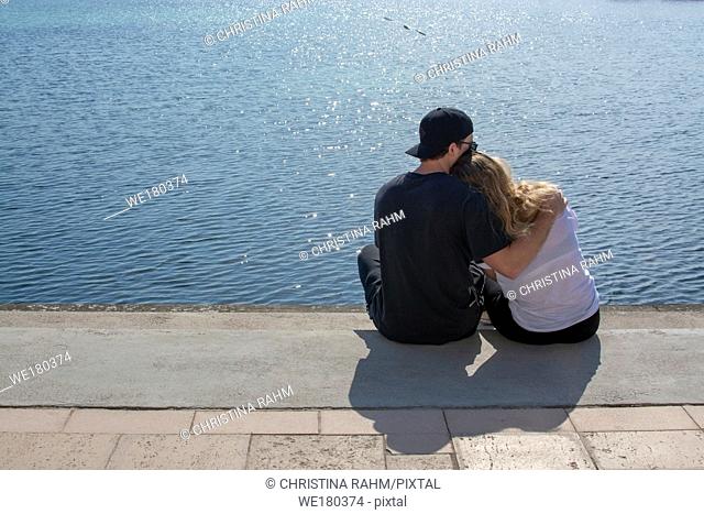 Young couple sit in sunshine watch water glitter on stone pier on a spring day in Mallorca, Spain